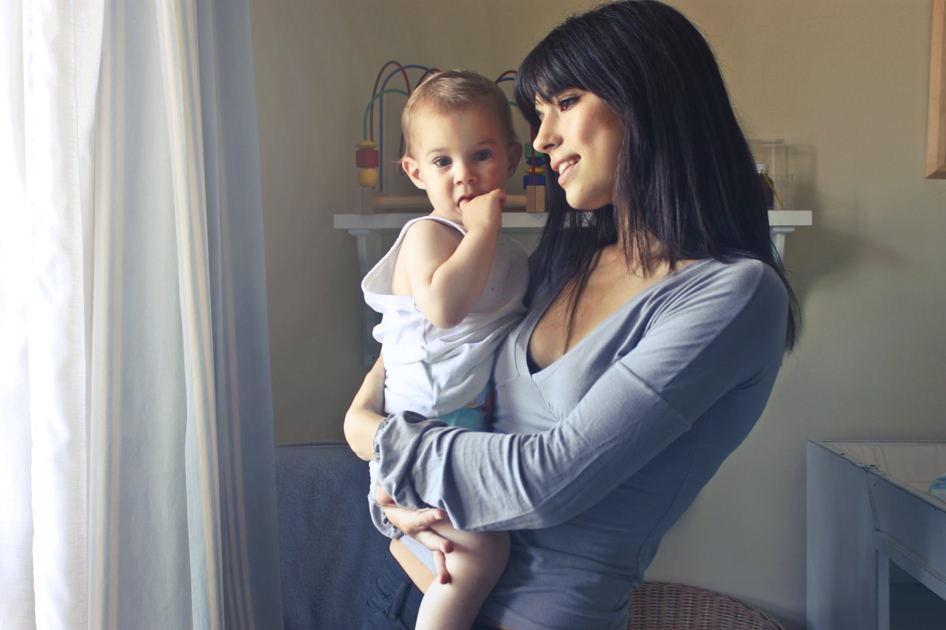 woman carrying baby boy wearing white tank top infront of white curtain inside the room